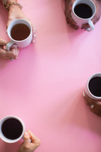 The hands of four women holding coffee cups on a pink table.