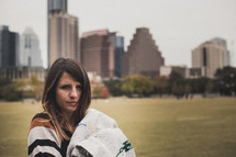 woman wrapped in a blanket standing in a park in a city 
