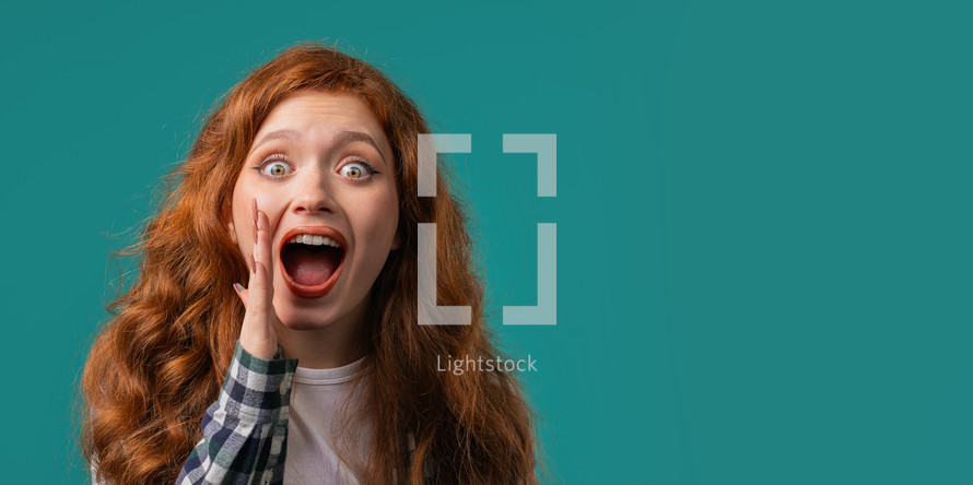 Attention, portrait of red haired young woman shouting, holding hand near open mouth isolated on blue background with copy space for your advertisement. High quality photo