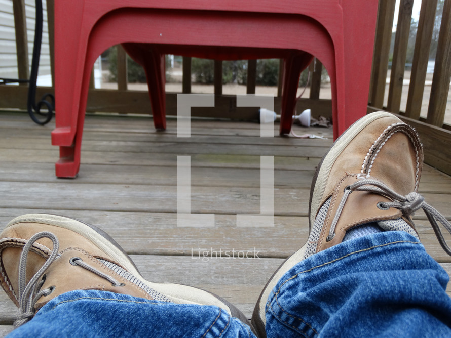 a person's legs laying on porch deck wearing jeans and brown boat shoes with red decor in background