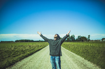 man standing in the middle of a dirt road with hands raised to God