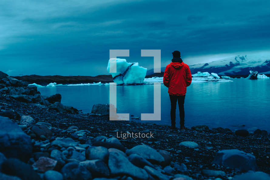 a man standing on a rocky shore looking out at ice and icebergs 