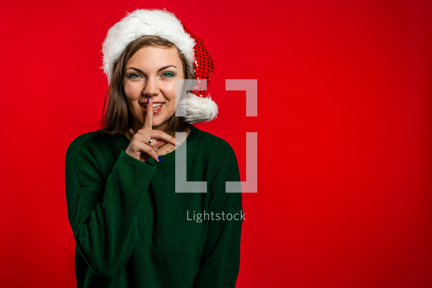 Beautiful woman in Christmas Santa hat holding finger on her lips over red