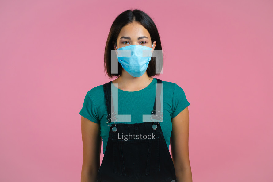 Young pretty girl in face medical mask during coronavirus pandemic. Portrait on pink background. Protection with respirator against COVID-19 outbreak.