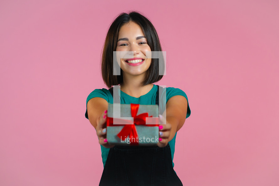 
Excited woman holding gift box and gives it by hands to camera on pink wall background. Girl smiling, she is happy with present. Studio portrait.