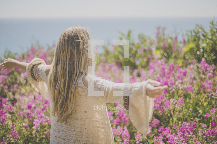 A blonde woman stands in a field of flowers with her arms outstretched.