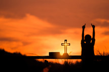 silhouette boy with hands raised and cross at sunset 
