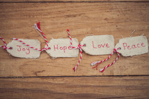 Christmas gift tags, reading "Joy," "Hope," "Love," and "Peace," lined up on a wood grain background.