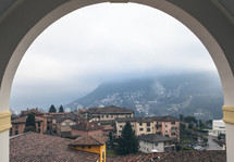 View through an arch of tile roofs, stucco buildings, and foggy mountains.