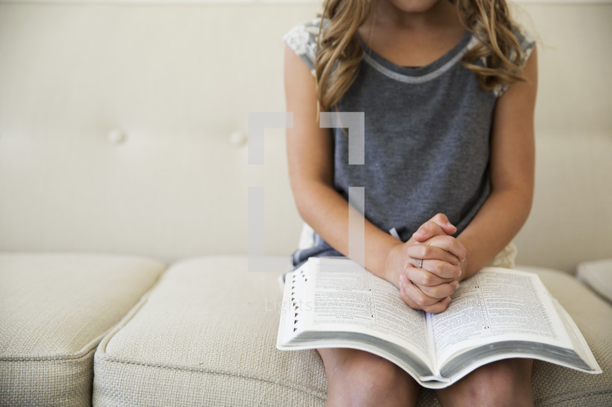 a girl child with praying hands over the pages of a Bible 