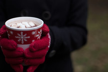 A Christmas cup of hot chocolate with marshmallows being held by a woman wearing red mittens.