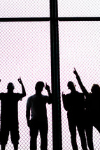 Silhouette of teen boys pointing up while standing behind a chain-link fence.