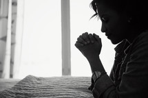 An African American woman kneeling in prayer beside her bed.