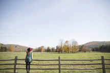 woman standing on a fence looking out at a field and mountain 