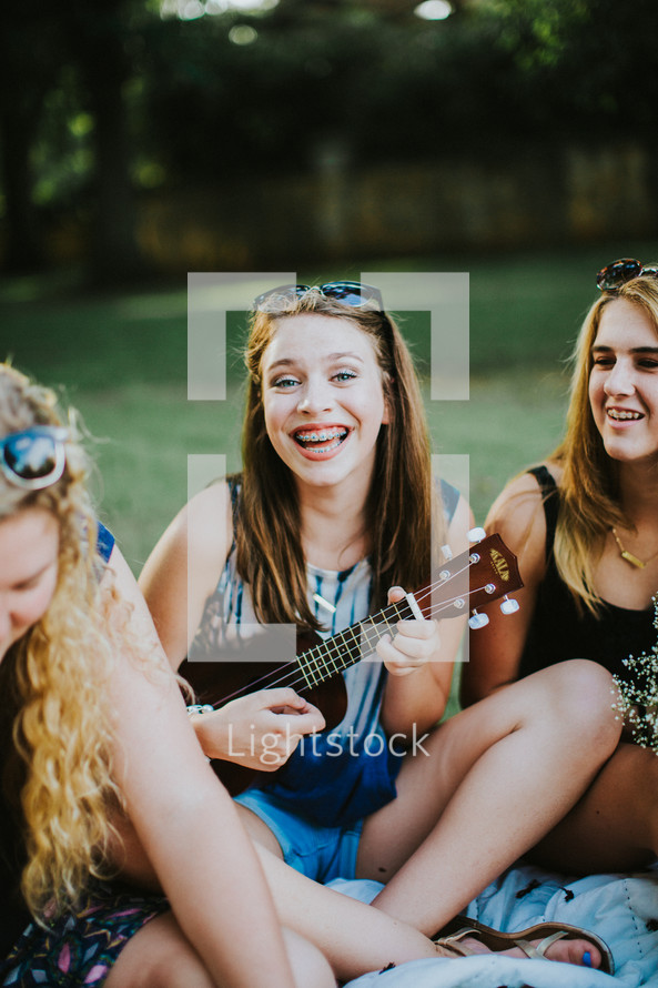 teen girls sitting on a blanket playing a ukulele and singing 