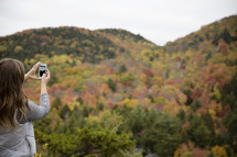 woman taking a picture with her phone of a fall mountain forest 