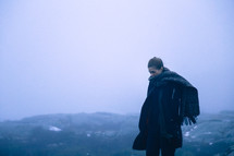 a woman in a coat and wrapped in a scarf standing on a mountaintop under an overcast sky 