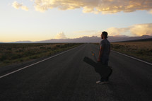 man standing in the middle of a road with a guitar case at sunset 