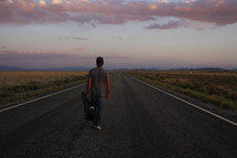 man standing in the middle of a road with a guitar case at sunset 