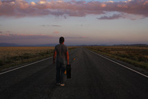 a man standing in the middle of a road with a suitcase at sunset 