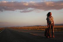 a young woman standing in the middle of a road with a suitcase at sunset 