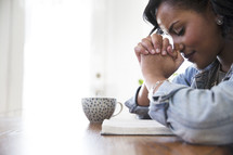 an African American woman sitting at a table reading a Bible and praying 