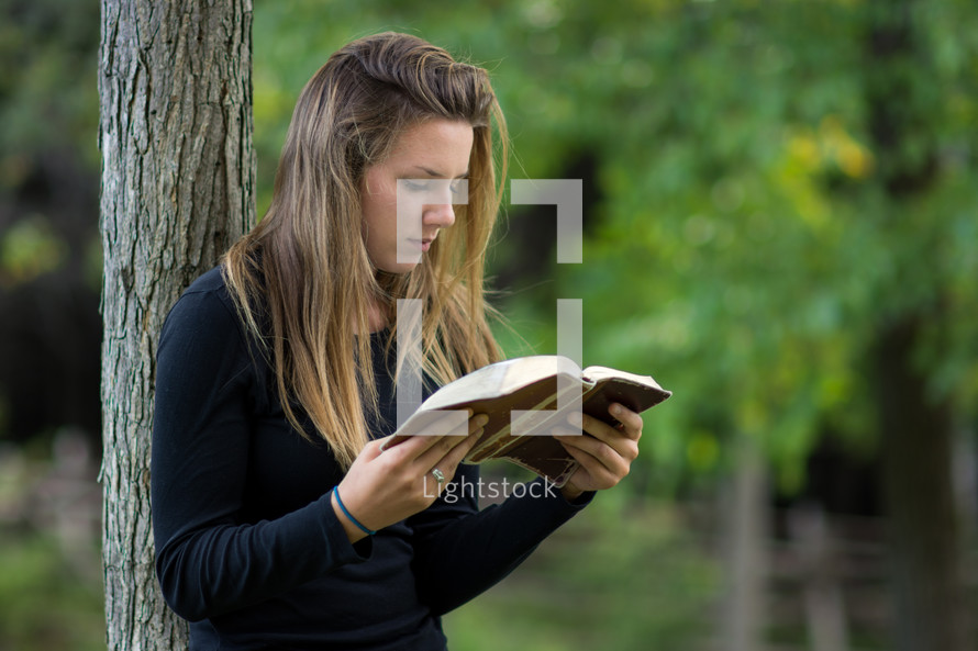 Girl standing outside, leaning against a tree, reading a bible.