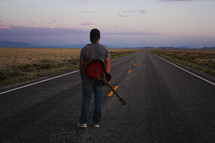 man standing in the middle of a road with a guitar at sunset 