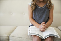 a girl child with praying hands over the pages of a Bible 