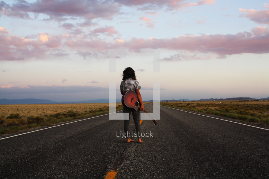 woman standing in the middle of a road with a guitar case at sunset 