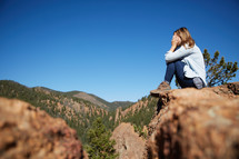 a woman covering her face sitting on the edge of a mountain 