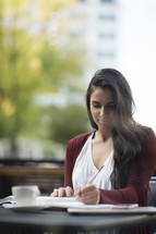 a young woman reading a Bible and writing in a journal at a table with a coffee mug and cellphone 