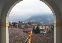 A view through an arch of tile roofs, stucco buildings and foggy mountains.
