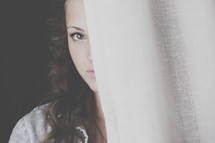 teenage girl timidly peeking from behind a curtain.