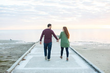 a couple holding hands walking on a beach pier