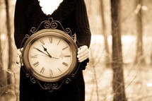 Woman in a black dress holding a large, ornate clock in a wooded area