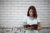 a young woman smiling as she reads a Bible in front of a white brick wall 