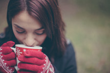 A young woman wearing red gloves drinks hot chocolate from a Christmas cup.