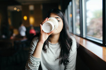 a young woman in a window seat of a coffee shop drinking coffee 