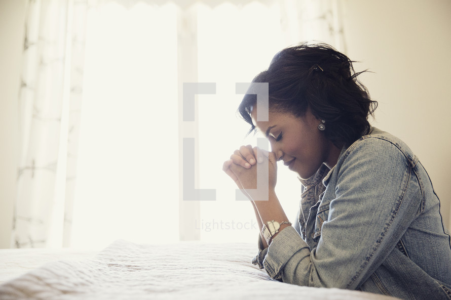 An African American woman kneeling in prayer beside her bed 