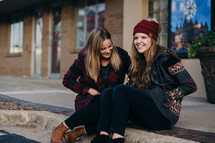young women in winter attire sitting on a curb talking 