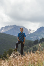 a young boy standing outdoors in front of a mountain peak 