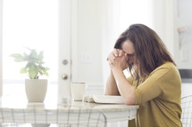 a woman praying over an open Bible sitting at a kitchen table 