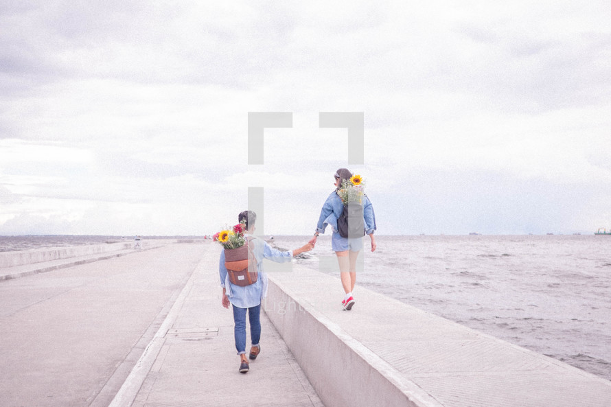 a couple on a date with flowers in their backpacks walking on a bridge over the ocean 