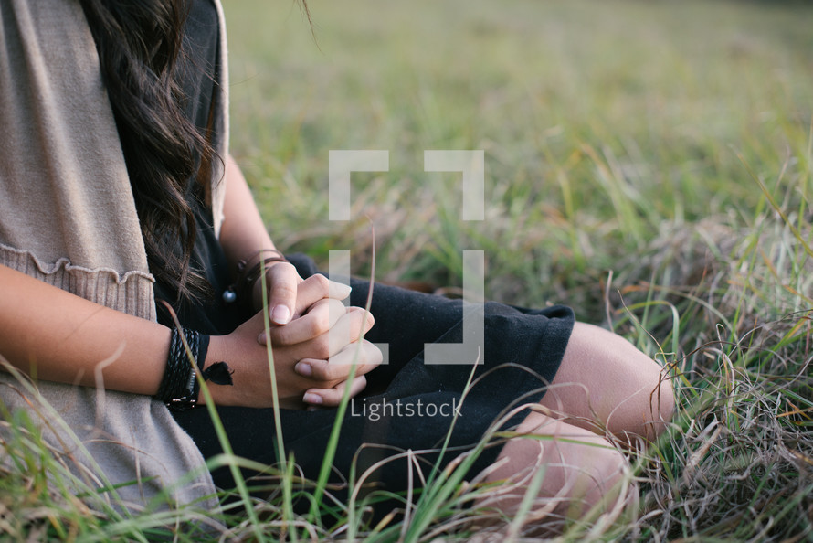 A young woman sitting in a the grass with her hands clasped.
