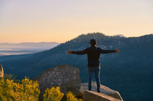 a man standing with open arms on a mountaintop looking out at the view 