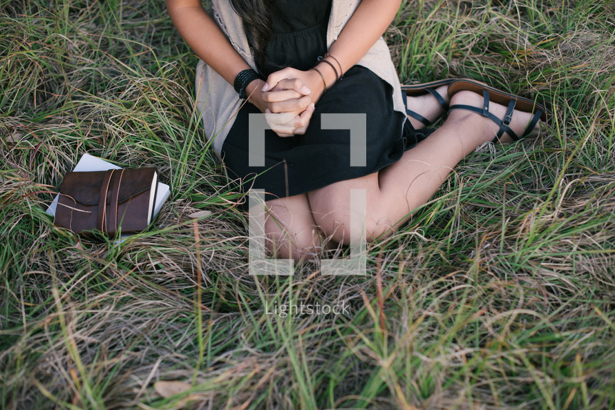A young girl sitting in grass with hands clasped.