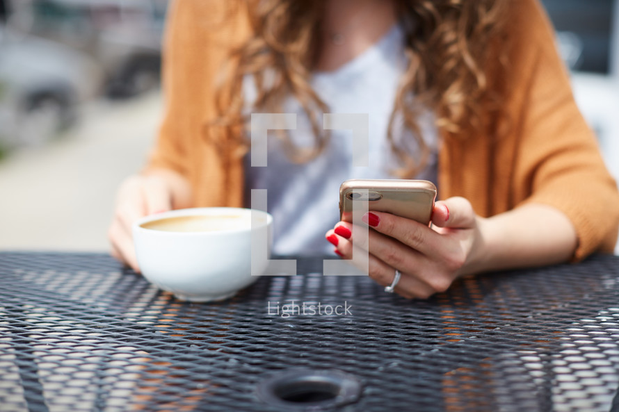a woman sitting at an outdoor table drinking coffee and looking at her cellphone 