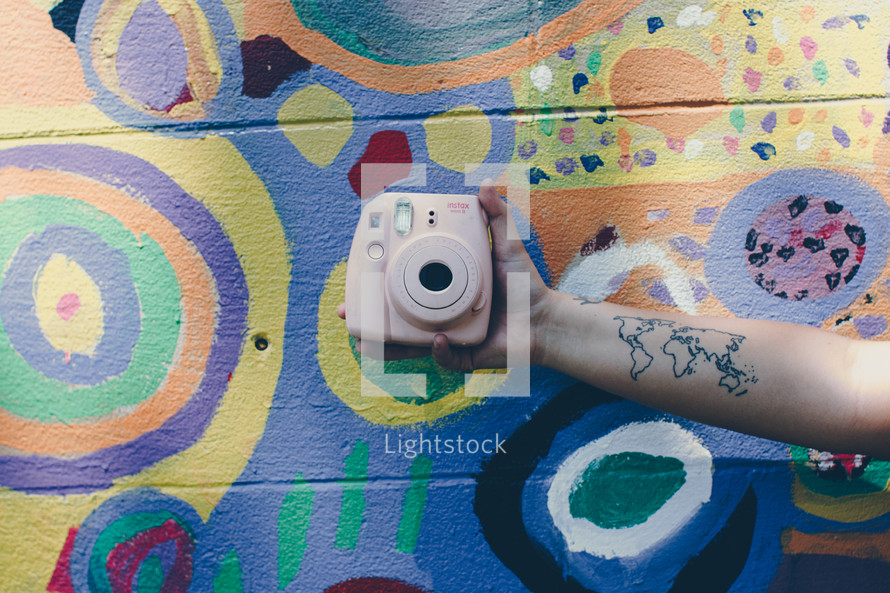 arm with a world map tattoo holding a camera in front of a painted wall 