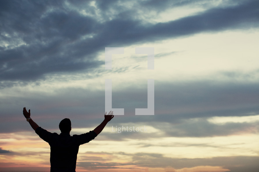 Silhouette of a man standing outside with arms raised in praise.